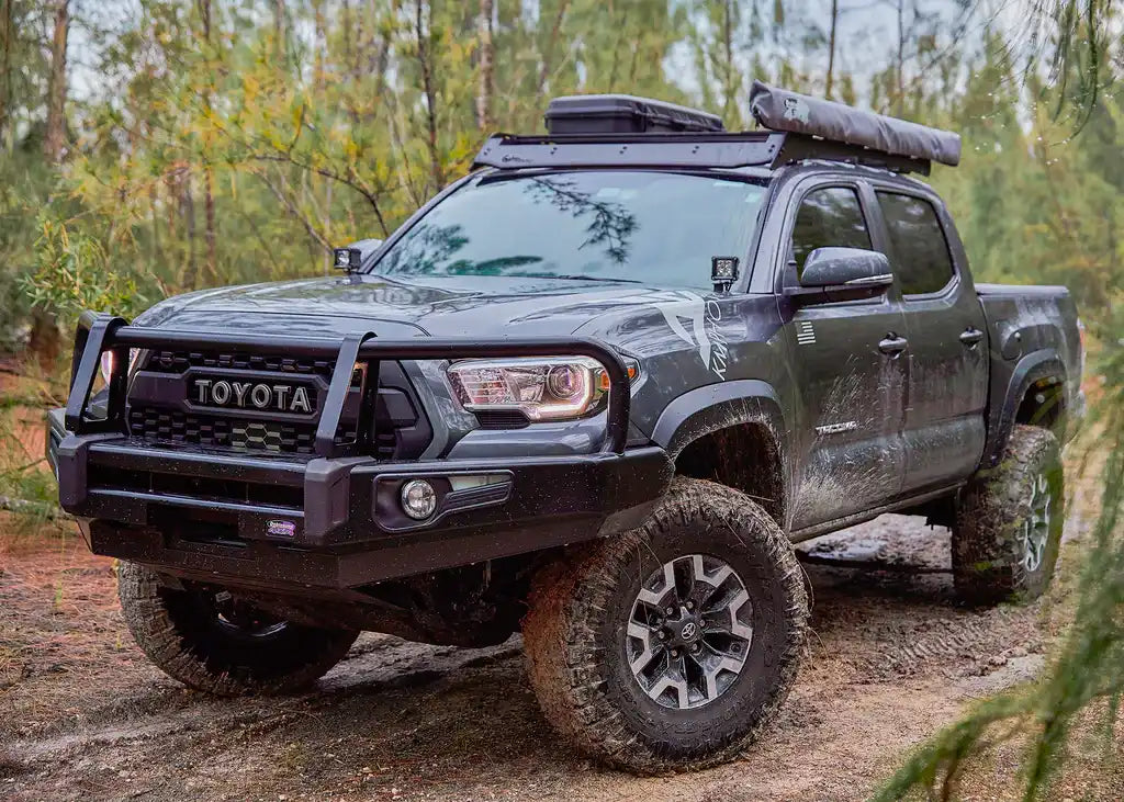 A dark grey Toyota Tacoma pickup truck, heavily modified for off-road use with a large front bumper and brush guard, lifted suspension, and knobby tires.