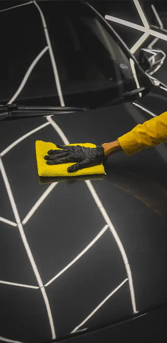 A person wearing a yellow long-sleeved shirt and black gloves wiping a black car hood with a yellow microfiber cloth.